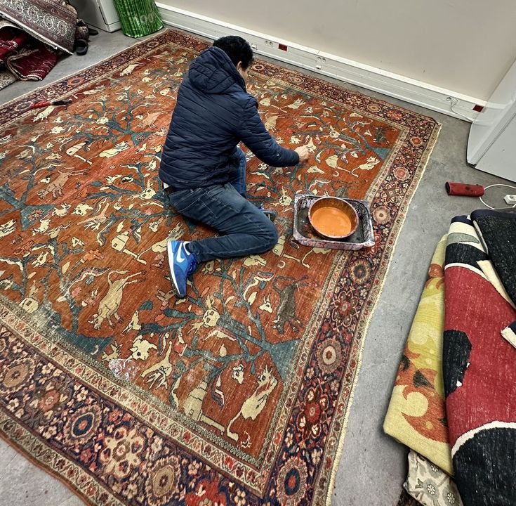 Artisan à genoux sur un grand tapis persan à décor animalier, repigmentant le champ rouge au pinceau